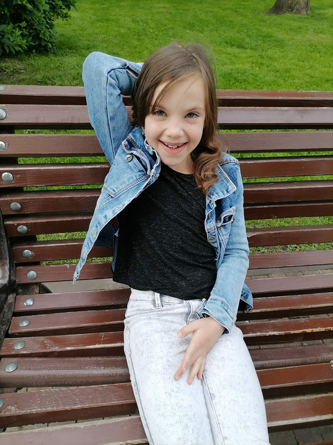 The girl sits on a bench in park. There is a flower ornament on her head